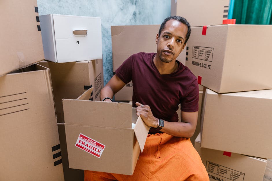 A man seated amid an arrangement of cardboard boxes inside a property, preparing for a home relocation. The boxes vary in size, some sealed with packing tape, and one features a red 'FRAGILE' sticker, indicating careful handling. The man, wearing a dark red T-shirt and orange pants, is looking directly at the camera while holding an open box. Behind him, additional boxes are stacked, with some placed on the floor and others on a surface. The environment is well-lit with natural or staged lighting. The image captures the process of packing and organizing belongings as part of furniture transport or a household move, illustrating the detailed loading process involved in removals services provided by Removal Companies Harrow. The setting outside of the boxes appears to be inside a residential property, emphasizing the packing phase of home relocation, reflecting professional removals procedures and logistics.