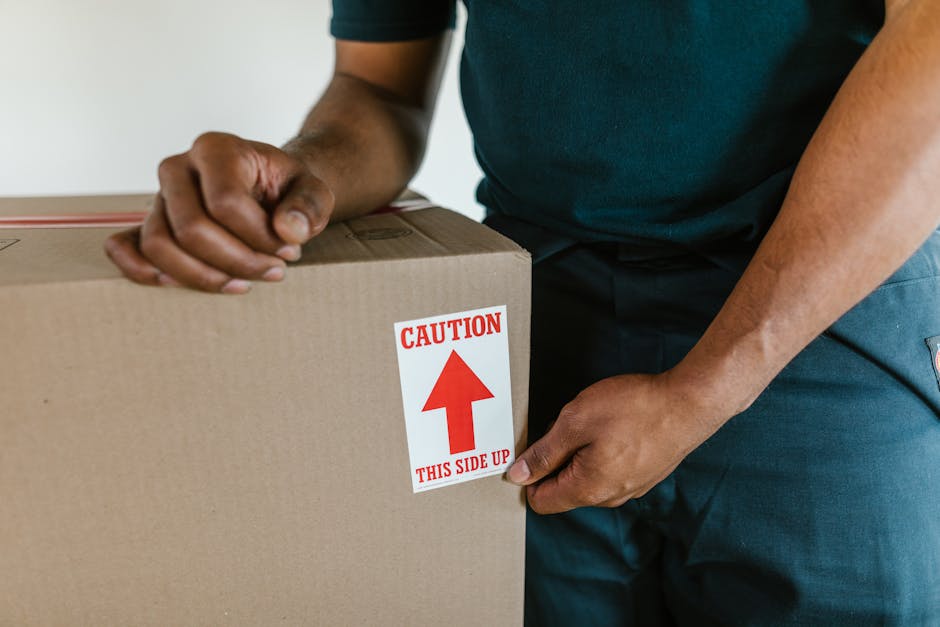 Close-up of a person wearing a dark green shirt and blue work trousers, securing a large cardboard box with a red and white sticker that reads 'CAUTION THIS SIDE UP' with an upward arrow. The person is holding the box firmly with both hands, one near the top and the other at the bottom, indicating careful handling during a home relocation process. The background features a neutral indoor setting, likely inside a property, with soft lighting illuminating the scene. This image captures the packing and handling phase typical of professional removals services, highlighting the importance of proper labelling for safe furniture transport. The scene reflects furniture packaging, the use of cardboard boxes, and cautious loading procedures associated with the moving process managed by Removal Companies Harrow, ensuring secure and organized home moves.