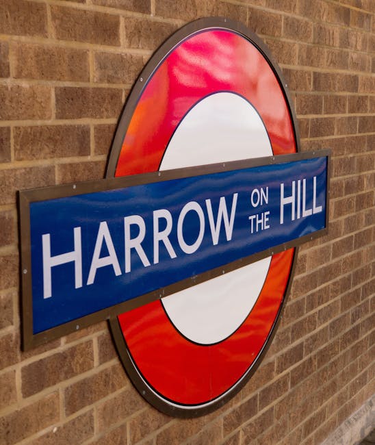 A large, circular London Underground sign with a red outer ring and a white inner circle, mounted on a brick wall interior. In front of the sign, a blue rectangular sign with white text reads 'HARROW ON THE HILL,' attached with five screws. The brick wall has a warm, reddish-brown tone, and the lighting appears to be indoor ambient light. This signage is in a setting that resembles a station or an area associated with movement and transport. The image subtly relates to home relocation and the logistics involved in furniture transport, which are key aspects of removals services offered by Removal Companies Harrow, especially when considering the risks of low quotes and the importance of professional moving assistance.