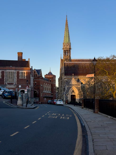 A street scene in a historic town with a narrow paved road marked 'BUS STOP' in white paint. On the left side, several parked cars are situated next to red-brick buildings with large windows and decorative detailing. On the right, a sidewalk runs alongside a tall, ornate church with a pointed green spire, stone archways, and intricate gothic-style architectural features. The church is partially shaded by surrounding trees, and a traditional street lamp is positioned near the sidewalk. The lighting suggests early morning or late afternoon. This setting illustrates a typical urban area where house removals or furniture transport might take place, with the church nearby indicating a residential and community locale. The image evokes a sense of the constraints and considerations associated with moving logistics in historic town centers, as handled by companies like Removal Companies Harrow.