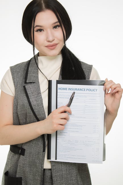 A professional moving company worker in a grey checkered vest and white shirt holds a clipboard with a home insurance policy document, using a pen to point at the form. The background shows a domestic interior with furniture covered in protective fabric and plastic wrapping, indicating ongoing packing and home relocation activities. Nearby, a moving trolley loaded with cardboard boxes, some secured with packing tape, is positioned on a wooden floor, reflecting the furniture transport process. The scene captures the preparation and logistics involved in house removals, illustrating the working environment during a typical furniture and belongings loading and moving operation, consistent with the services offered by Removal Companies Harrow in the context of professional removals and packing services.
