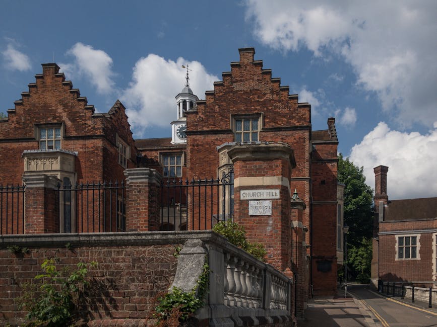 Exterior view of a historic brick building on Church Hill, with a decorative stone balustrade in the foreground and a black wrought iron fence along the top of the brick wall. The building features ornate brickwork with stepped gables, a small tower with a clock and weather vane, and large windows with cream-colored frames. A narrow street runs alongside the building, partially visible on the right, with a sidewalk and some greenery. The sky above is partly cloudy, casting natural light on the scene, which depicts the type of location where house removals or furniture transport services by Removal Companies Harrow may take place, especially during home relocation or moving activities involving careful planning of street loading permits.