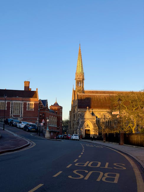 The image shows a quiet street scene during daylight hours in a historic town, featuring a prominent stone church with a tall, pointed spire and detailed architectural features, positioned on the right side of the street. The church is surrounded by traditional red-brick buildings, some with chimney stacks, which are located on the left side of the road. Several parked cars are lined along the curb close to the buildings. The street curves gently to the left, with a clearly visible yellow 'BUS STOP' marking on the asphalt and a dashed double yellow line along the edge of the road. A concrete pavement runs parallel to the street, leading up to the entrances of the buildings and the church. The scene is illuminated with warm, natural sunlight, casting shadows and highlighting the textures of the stone and brickwork. This setting may relate to house removals or local transport logistics, as seen in services provided by Removal Companies Harrow, who facilitate furniture transport and packing during home relocation processes.