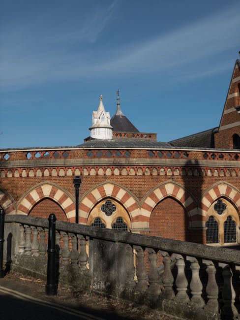 The image depicts a historic brick building with a rounded, parapet wall featuring decorative arches and circular window openings. The building has a conical roof with a small spire and is topped with ornate white cupolas. In the foreground, a low stone or concrete balustrade with black metal posts runs along a pavement or pathway, casting shadows on the ground. The scene is illuminated by bright natural daylight with a clear blue sky overhead, highlighting the architectural details. This setting is typical of a heritage or ecclesiastical structure, relevant to house removals and moving services provided by Removal Companies Harrow, especially in the context of home relocation or transporting valuable or delicate furnishings within a historic environment.
