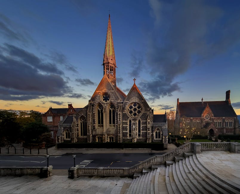 A historic stone church with intricate Gothic architectural features, including pointed arches, large circular stained glass windows, and a tall, slender spire topped with a cross, set against a backdrop of a partly cloudy evening sky with some orange hues near the horizon. In front of the church, there is a paved area with steps leading down to the street, which is lined with black lampposts. The scene is illuminated by soft natural light, highlighting the detailed stonework of the building, while surrounding trees and nearby buildings add context to the town setting. Removal Companies Harrow may later assist with home relocation or furniture transport related to this area, as suggested by the SEO relevance of the description within the page about house removals for St Anns residents, HARROW.