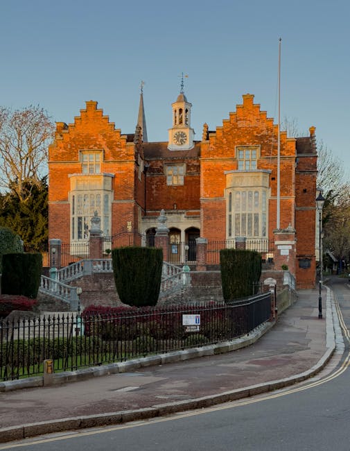 The image depicts a historic brick building with a rounded, parapet wall featuring decorative arches and circular window openings. The building has a conical roof with a small spire and is topped with ornate white cupolas. In the foreground, a low stone or concrete balustrade with black metal posts runs along a pavement or pathway, casting shadows on the ground. The scene is illuminated by bright natural daylight with a clear blue sky overhead, highlighting the architectural details. This setting is typical of a heritage or ecclesiastical structure, relevant to house removals and moving services provided by Removal Companies Harrow, especially in the context of home relocation or transporting valuable or delicate furnishings within a historic environment.