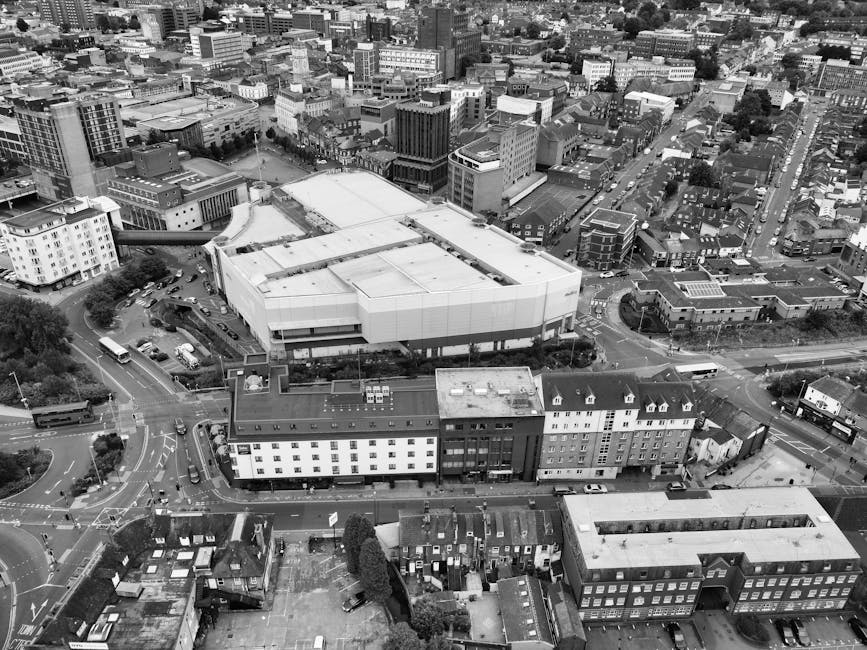 An aerial view in black and white showing a dense cityscape with a mix of modern and older buildings. In the foreground, there is a large, multi-story building with a flat roof, surrounded by smaller structures including rectangular residential and commercial properties with pitched roofs, some featuring chimneys, and several with rooftop access areas. A network of roads and roundabouts is visible, with cars, buses, and trucks moving through the area. Adjacent to the large building, there is a parking lot with parked vehicles and a few trees. The background reveals more high-rise and mid-rise buildings, indicating a busy urban environment. The scene captures the dense infrastructure and various urban elements typical of a city centre, illustrating the logistics involved in house removals and furniture transport within an urban setting, as conducted by Removal Companies Harrow during a home relocation or office move.