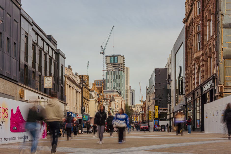 A busy city centre street scene during daytime shows pedestrians walking along the pavement, some carrying shopping bags or backpacks. On the sides of the street are retail stores with large storefront windows, including a shop with a pink advertisement on the left. The buildings are a mix of modern and historic architecture, with a construction crane and a tall building under construction visible in the background. The sky is overcast, casting diffuse lighting across the scene. Although no items from a house removal process are visible, this urban environment reflects the logistical context of moving or relocation services such as those provided by Removal Companies Harrow, especially when coordinating furniture transport or home relocation in a bustling commercial area.