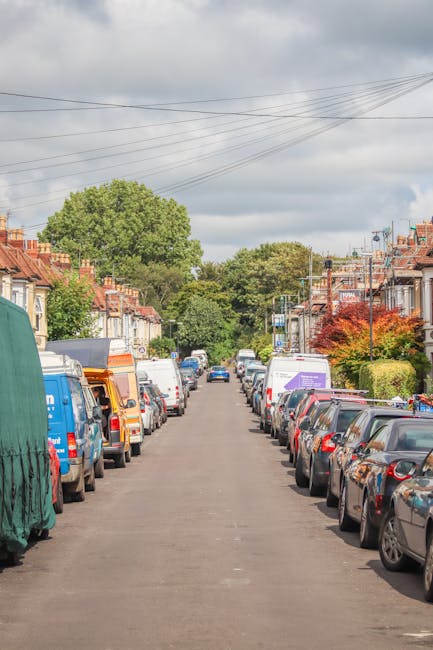 A residential street lined with parked cars on both sides, including various models of vans, some with covered loads or open doors revealing packing materials and furniture being prepared for removal. The pavement runs straight between rows of detached and semi-detached houses with brick exteriors, small front gardens, and diverse trees and bushes providing greenery along the street. Overhead, power lines stretch across a cloudy sky, creating a network above the scene. In the background, larger trees are visible, indicating a suburban area. The image captures the typical environment encountered during house removals and furniture transport, as professional moving services like Removal Companies Harrow coordinate the loading process, utilizing trolleys and packing materials such as cardboard boxes, bubble wrap, and blankets to ensure safe transportation of belongings. The overall scene reflects an active home relocation setting with vehicles positioned for optimal loading and unloading.