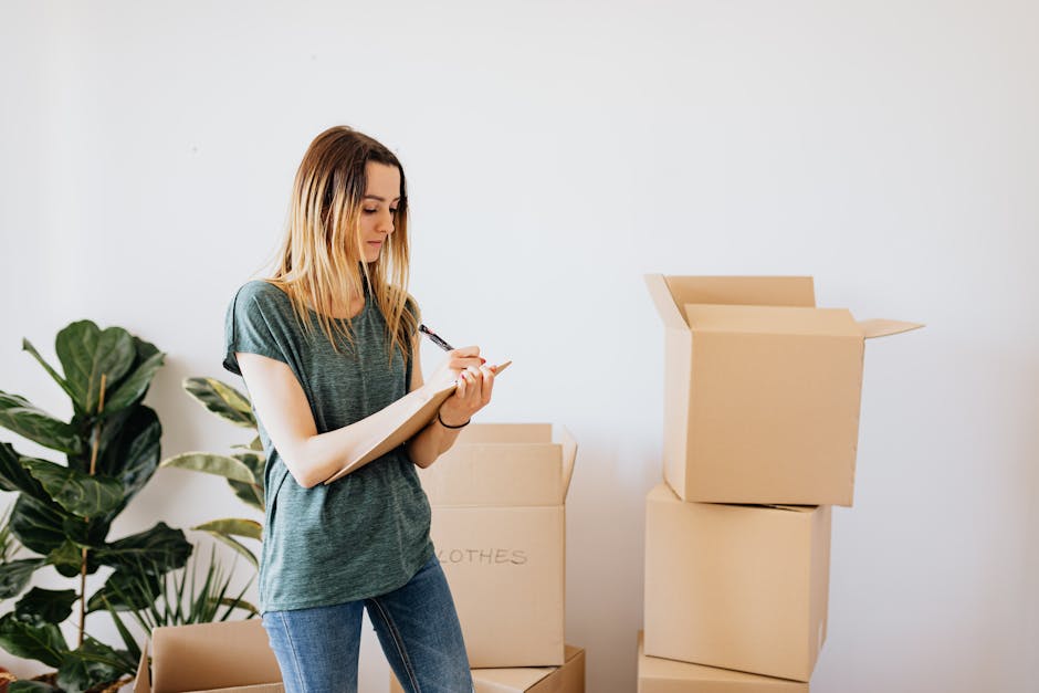 A young woman with long blonde hair wearing a casual grey t-shirt and blue jeans is inside a residential setting, surrounded by several cardboard boxes used for packing belongings during a home relocation. She appears to be taking notes or making a checklist on a notepad with a pen, as part of the packing and moving process. Behind her, there is a large leafy green indoor plant, and the boxes are stacked in an orderly manner, some open to reveal packed items. The room has a plain white wall, and natural daylight illuminates the scene, emphasizing the organized environment associated with furniture transport and professional removals services. This image relates to the activities involved in house removals, highlighting aspects of packing, planning, and preparation for a house move, which is part of the services offered by Removal Companies Harrow in the context of their HA3 price checklist for home relocation purposes.