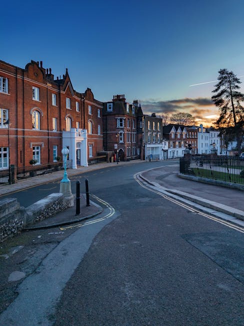 A quiet residential street during sunset featuring a row of red-brick and white-painted terraced houses with bay windows and chimneys. The street curves gently to the right, with a paved sidewalk and black iron railings lining a small grassy area on the right. On the left, there is a street lamp with a blue-painted base, and a few black bollards and a concrete barrier near the curb. The sky is clear with a warm, orange glow near the horizon, casting soft light over the scene. The area appears calm and suitable for home relocations or furniture transport, as part of a house removal process, with no moving activity visible but indicating an environment where logistics and moving services could be performed by professionals like Removal Companies Harrow.