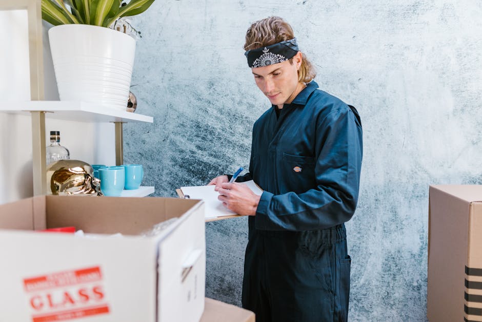 A young woman with long blonde hair wearing a casual grey t-shirt and blue jeans is inside a residential setting, surrounded by several cardboard boxes used for packing belongings during a home relocation. She appears to be taking notes or making a checklist on a notepad with a pen, as part of the packing and moving process. Behind her, there is a large leafy green indoor plant, and the boxes are stacked in an orderly manner, some open to reveal packed items. The room has a plain white wall, and natural daylight illuminates the scene, emphasizing the organized environment associated with furniture transport and professional removals services. This image relates to the activities involved in house removals, highlighting aspects of packing, planning, and preparation for a house move, which is part of the services offered by Removal Companies Harrow in the context of their HA3 price checklist for home relocation purposes.