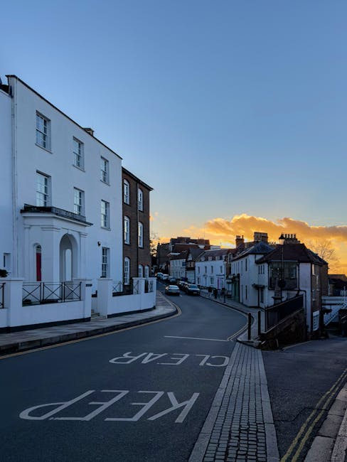 A quiet residential street in Wealdstone during sunset, with a row of terraced and semi-detached houses lining the curved road. The houses feature a mix of white and brick facades, some with small front gardens and railings. The road has clear white markings including a 'KEEP LEFT' instruction painted on the tarmac, and the pavement is composed of paving slabs with tactile strips near the corner for pedestrian access. A black street lamp is visible on the sidewalk, and a few parked cars can be seen further down the street. The sky is painted with warm orange and yellow hues from the setting sun, casting long shadows across the scene. This location exemplifies a typical suburban area suitable for house removals and home relocation activities. The image might be used to support content related to house removals, moving logistics, or packing and transport services provided by Removal Companies Harrow, especially relevant for same-day moves in Harrow.