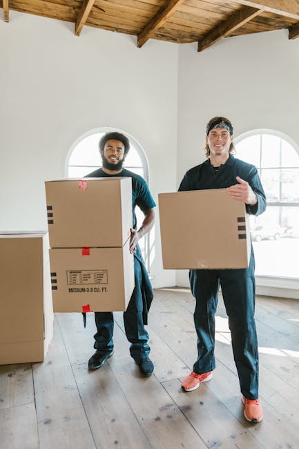 Two movers from Removal Companies Harrow are inside a well-lit room with white walls, carrying medium-sized cardboard boxes. One mover, on the left, is holding two boxes stacked atop each other with red and black tape securing them, while the other mover, on the right, is holding a single box at waist height with a smile, wearing casual dark clothing and red sneakers. The room features a wooden ceiling with visible beams and natural light coming through two arched windows behind them. The scene captures the process of home relocation, where the movers are preparing to load the boxes onto a moving van or transition them through the space. Packaging materials, including cardboard, are visible, emphasizing packing and moving activities associated with home furniture transport and furniture transport logistics, typical of professional removal services. The environment appears organized, and the movers are engaged in a careful loading process in preparation for a same-day move in Wealdstone, Harrow.