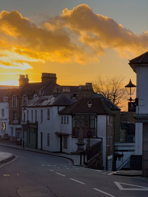 A quiet residential street in Wealdstone during sunset, with a row of terraced and semi-detached houses lining the curved road. The houses feature a mix of white and brick facades, some with small front gardens and railings. The road has clear white markings including a 'KEEP LEFT' instruction painted on the tarmac, and the pavement is composed of paving slabs with tactile strips near the corner for pedestrian access. A black street lamp is visible on the sidewalk, and a few parked cars can be seen further down the street. The sky is painted with warm orange and yellow hues from the setting sun, casting long shadows across the scene. This location exemplifies a typical suburban area suitable for house removals and home relocation activities. The image might be used to support content related to house removals, moving logistics, or packing and transport services provided by Removal Companies Harrow, especially relevant for same-day moves in Harrow.